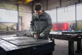 A person wearing safety equipment uses an angle grinder on a metal worktable.
