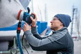A person wearing safety equipment sands a boat hull with a random orbit sander.