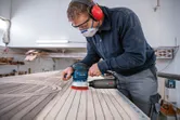 A person wearing safety equipment sands wooden planks with a random orbit sander.