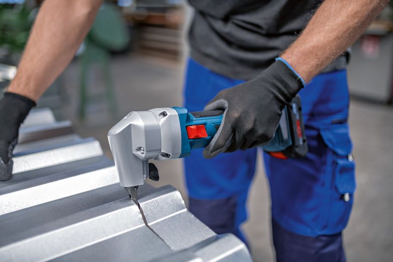 A person wearing safety equipment cuts sheet metal using a cordless nibbler.