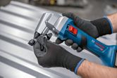 A person wearing safety equipment uses a cordless nibbler on a metal sheet.