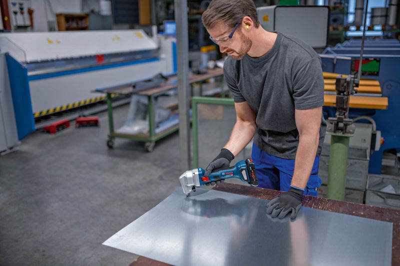 A person wearing safety equipment uses a cordless nibbler to cut sheet metal.