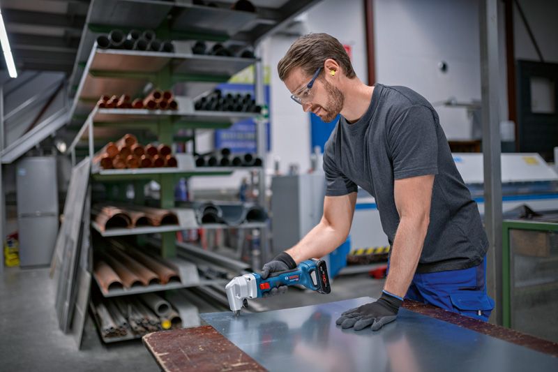 A person wearing safety equipment cuts sheet metal with a cordless nibbler.
