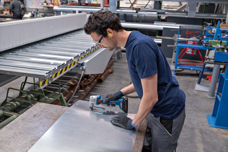 A person wearing safety equipment cuts sheet metal with a cordless metal shear.