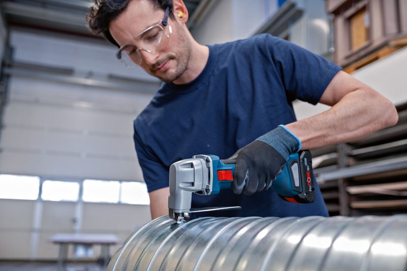 A person wearing safety equipment cuts a metal duct with a cordless metal shear.