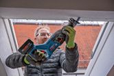 A person wearing safety equipment uses a reciprocating saw to cut into a window frame.