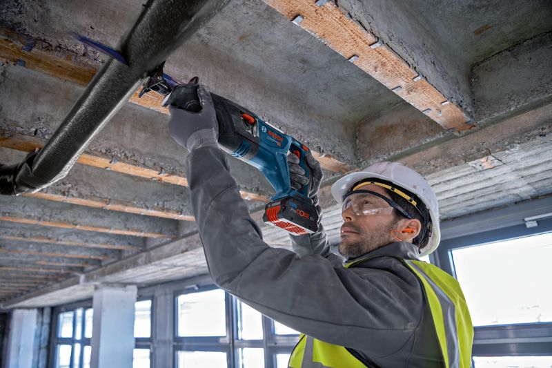A person wearing safety equipment uses a reciprocating saw to cut a pipe overhead.