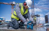 A person wearing safety equipment uses a cordless angle grinder on a concrete surface.