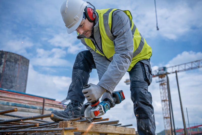 A person wearing safety equipment uses a cordless angle grinder to cut rebar.