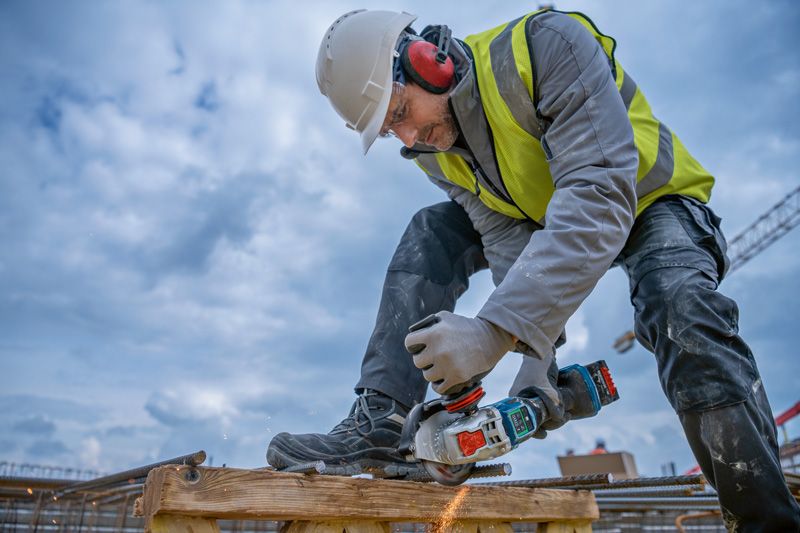 A person wearing safety equipment cuts rebar with a cordless angle grinder.