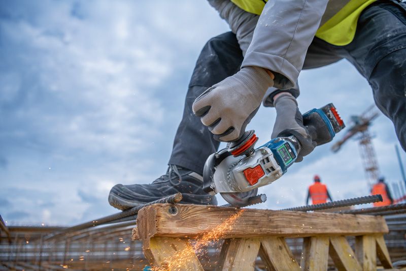 A person wearing safety equipment grinds rebar with a cordless angle grinder on a construction site.