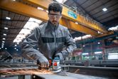 A person wearing safety equipment grinds metal with a cordless angle grinder in a factory.