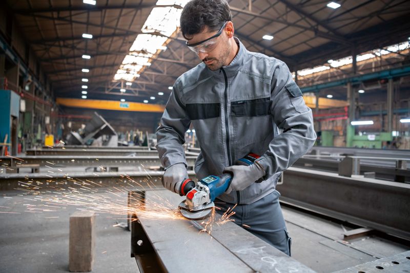 A person wearing safety equipment grinds a metal beam with a cordless angle grinder.