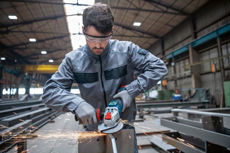 A person wearing safety equipment grinds metal with a cordless angle grinder in a factory.