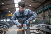 A person wearing safety equipment grinds metal with a cordless angle grinder.