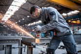 A person wearing safety equipment grinds metal with a cordless angle grinder in a factory.