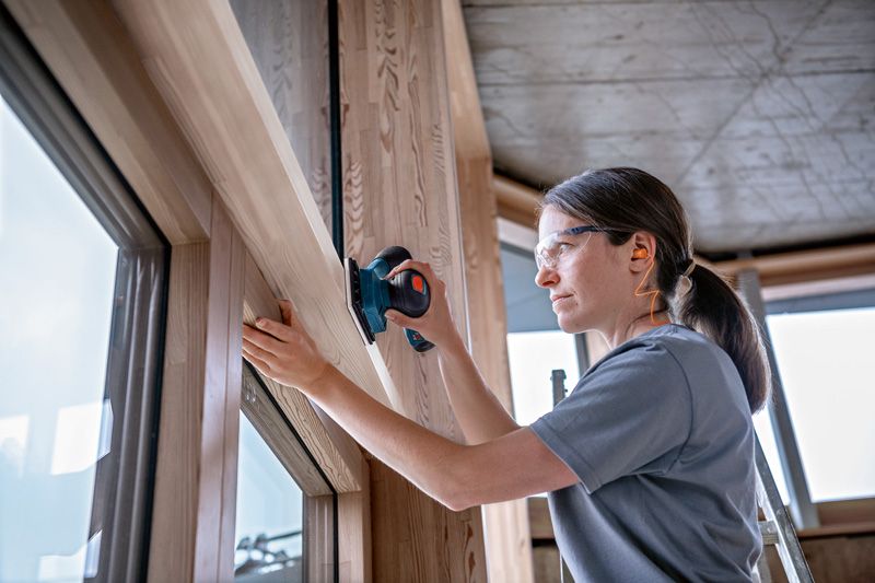A person wearing safety equipment sands a wooden window frame with a cordless orbital sander.