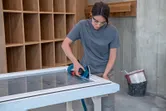 A person wearing safety equipment sands a window frame with a cordless orbital sander.