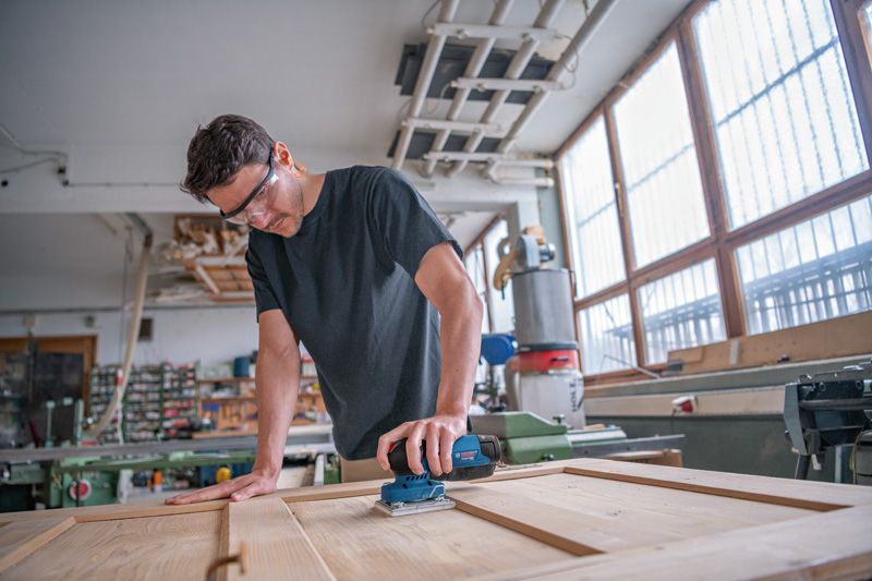 Person wearing safety equipment sands a wooden door with a cordless orbital sander.