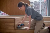 A person wearing safety equipment sands wooden stairs with a cordless orbital sander.