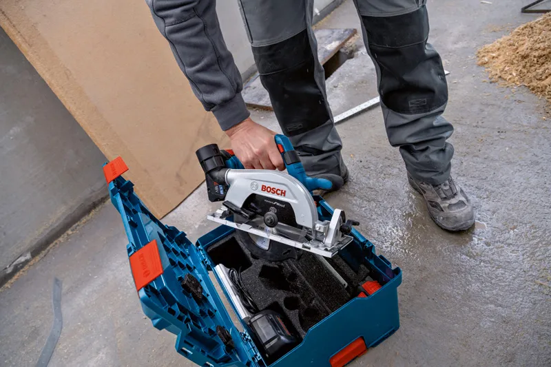 A person wearing safety equipment lifts a circular saw from a tool kit on a workshop floor.