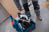 A person wearing safety equipment lifts a circular saw from a tool kit on a workshop floor.
