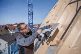 Person wearing safety equipment cuts wooden boards on a slanted roof with a circular saw.