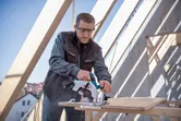 A person wearing safety equipment uses a circular saw to cut plywood on a construction site.