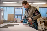 A person wearing safety equipment sands a wooden surface with a random orbit sander.