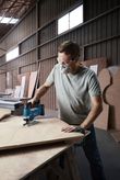 A person wearing safety equipment uses a jigsaw to cut curved lines in plywood.