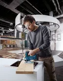 A person wearing safety equipment cuts wood with a jigsaw in an indoor construction area.