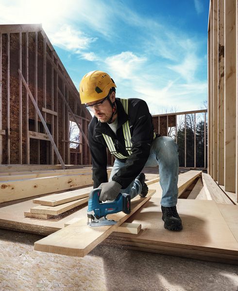 A person wearing safety equipment uses a jigsaw to cut wood at a construction site.