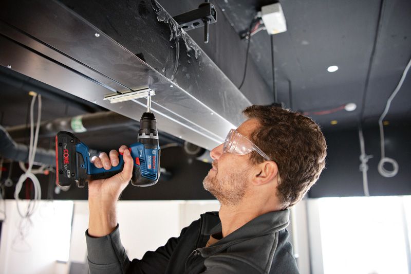 GSB 18V-55 A person wearing safety equipment uses a cordless combi drill to drive a screw overhead.