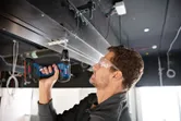 A person wearing safety equipment uses a cordless combi drill to drive a screw overhead.