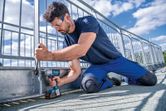 A person wearing safety equipment fastens a metal fence with a cordless impact wrench.