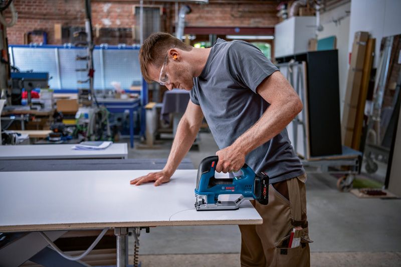 A person wearing safety equipment uses a cordless jigsaw to cut a precise curve in a panel.
