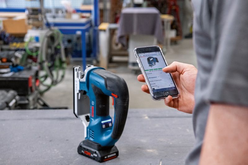 A person uses a smartphone to adjust a cordless jigsaw in a workshop.
