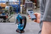 A person uses a smartphone to adjust a cordless jigsaw in a workshop.
