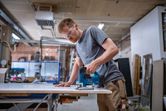 A person wearing safety equipment cuts wood with a cordless jigsaw in a workshop.