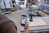 A person uses a smartphone to reference a cordless drill in a woodworking shop.