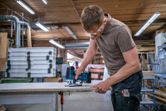 A person wearing safety equipment cuts a wood board with a jigsaw in a workshop.