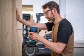 A person wearing safety equipment uses a cordless drill to drive a screw into plywood.