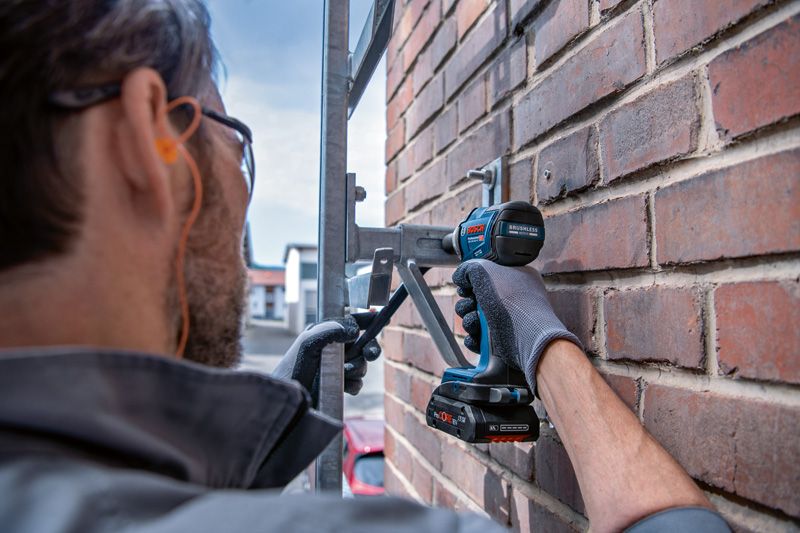 Person wearing safety equipment uses a power drill to fasten a metal bracket to a brick wall.