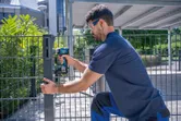 A person wearing safety equipment uses a cordless drill to fasten a metal fence post.