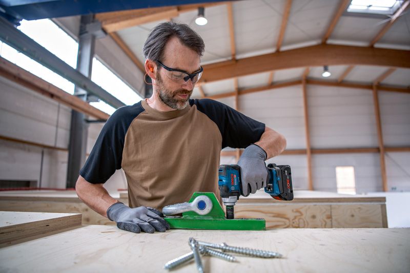 GDS 18V-330 HC A person wearing safety equipment uses a cordless drill on a wooden workbench.