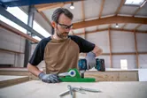A person wearing safety equipment uses a cordless drill on a wooden workbench.