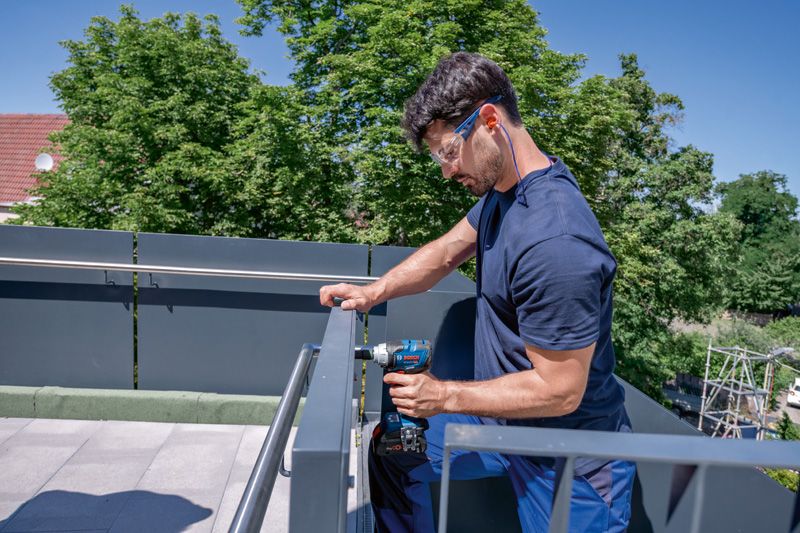GDS 18V-330 HC A person wearing safety equipment tightens a screw on a metal railing with a power drill.