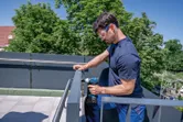 A person wearing safety equipment tightens a screw on a metal railing with a power drill.