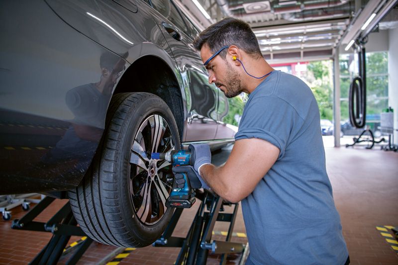 A person wearing safety equipment uses an impact wrench to tighten bolts on a car wheel.