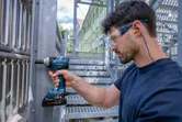 A person wearing safety equipment uses a cordless impact wrench on a metal structure.